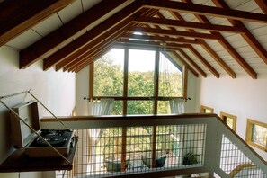 A room with a high ceiling, wooden beams, a large window, and a view of the outdoors. - Sycamore Treehouse at The Meadow by HoneyTree (Fredericksburg)