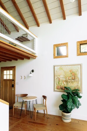 Une pièce avec un plancher en bois, une table à manger avec des chaises, une grande plante en pot et un tableau accroché au mur.