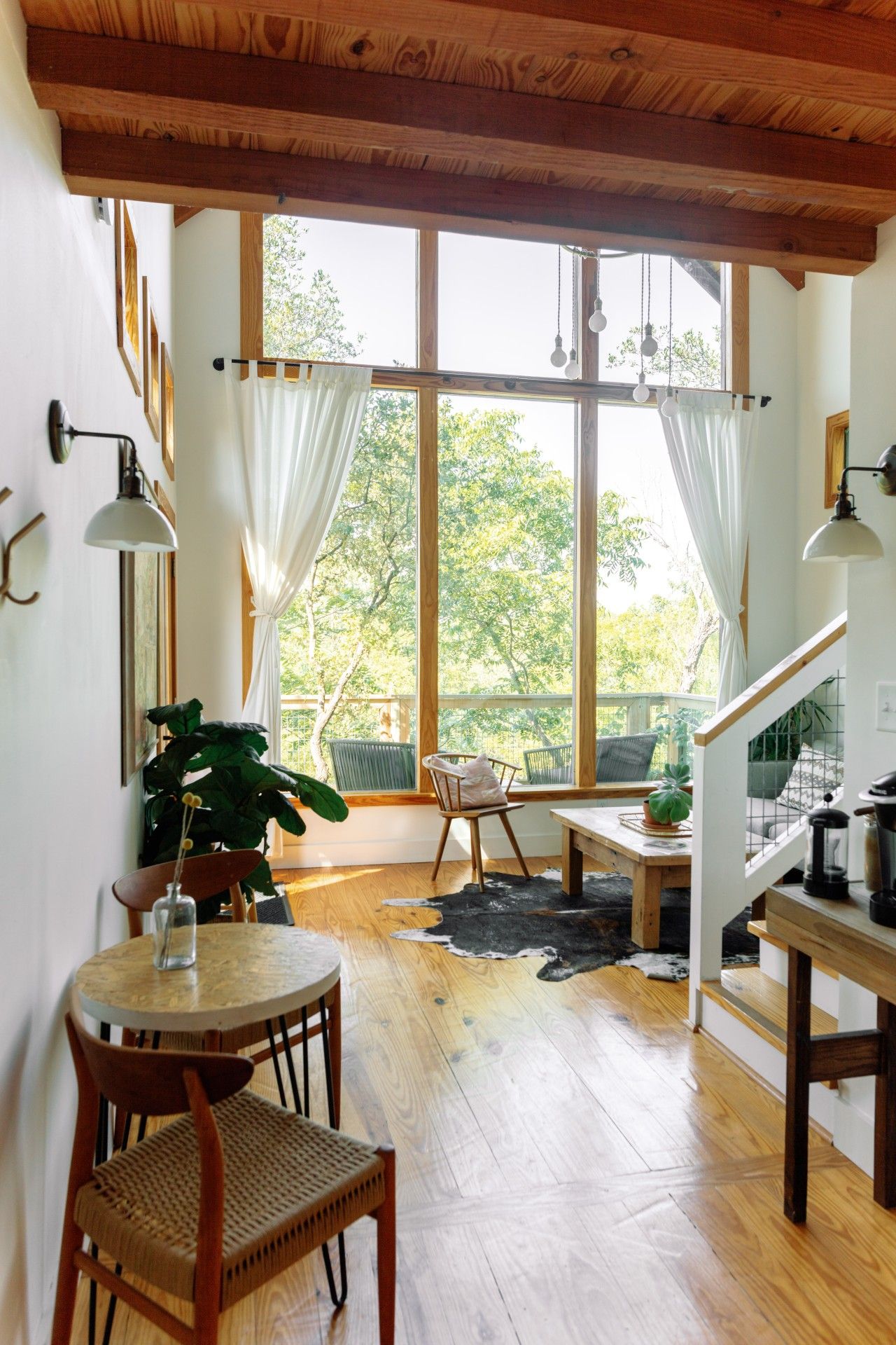 A cozy interior with a dining area, wooden flooring, and large windows offering a view of greenery.