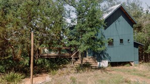 A rustic cabin with a wooden deck and a metal roof, surrounded by trees and vegetation. - Sycamore Treehouse at The Meadow by HoneyTree (Fredericksburg)