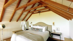 A bedroom with a sloped ceiling, a bed with a beige headboard, bedside tables, a floor lamp, and a robe hanging on a hook. - Sycamore Treehouse at The Meadow by HoneyTree (Fredericksburg)