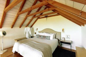 Une chambre avec un plafond en pente, un lit doté d’une tête de lit beige, des tables de chevet, une lampe sur pied et un peignoir suspendu à un crochet.