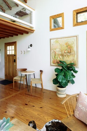 A room with a wooden floor, a dining table with chairs, a large potted plant, and framed artwork on the wall. - Sycamore Treehouse at The Meadow by HoneyTree (Fredericksburg)