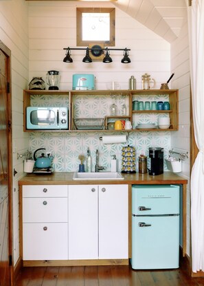 A compact kitchen with a vintage microwave, a small refrigerator, and wooden shelves above the counter. - Live Oak Treehouse at The Meadow (Fredericksburg)