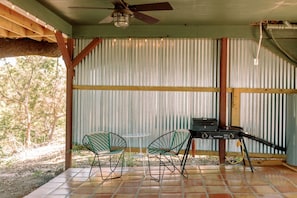 A covered outdoor area with a ceiling fan, two chairs, a grill, and a corrugated metal wall.