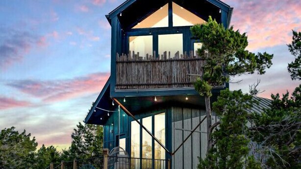 A two-story wooden cabin with a balcony, surrounded by trees and a wooden deck.