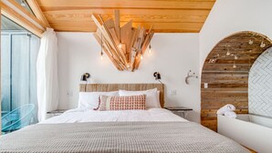 A modern bedroom with a wooden ceiling, a large bed with white bedding, a blue chair, and a wooden wall with a herringbone pattern.