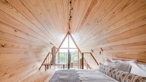 A wooden cabin bedroom with a bed, pillows, and a view through a triangular window. - Blue Sage No. 2 - The Floating A-Frame (Fredericksburg)