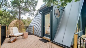 A wooden deck with a wicker chair and a small table, a door with a circular window, and a metal railing. - Blue Sage No. 2 - The Floating A-Frame (Fredericksburg)