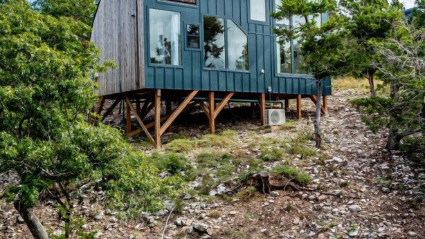 Eine moderne Holzhütte mit großen Fenstern, auf Stelzen erhöht, umgeben von Bäumen und felsigem Gelände.