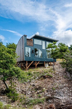 A modern wooden cabin with large windows, elevated on stilts, surrounded by trees and rocky terrain.