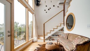 Amplia sala de estar con una escalera de madera, un sofá, un espejo redondo y vistas al exterior gracias a amplios ventanales.