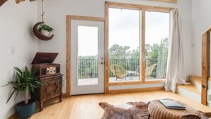 Una habitación con suelo de madera, un amplio ventanal corredera que da a un balcón, un aparador de madera rústica, una planta colgante y un otoman de cuero.