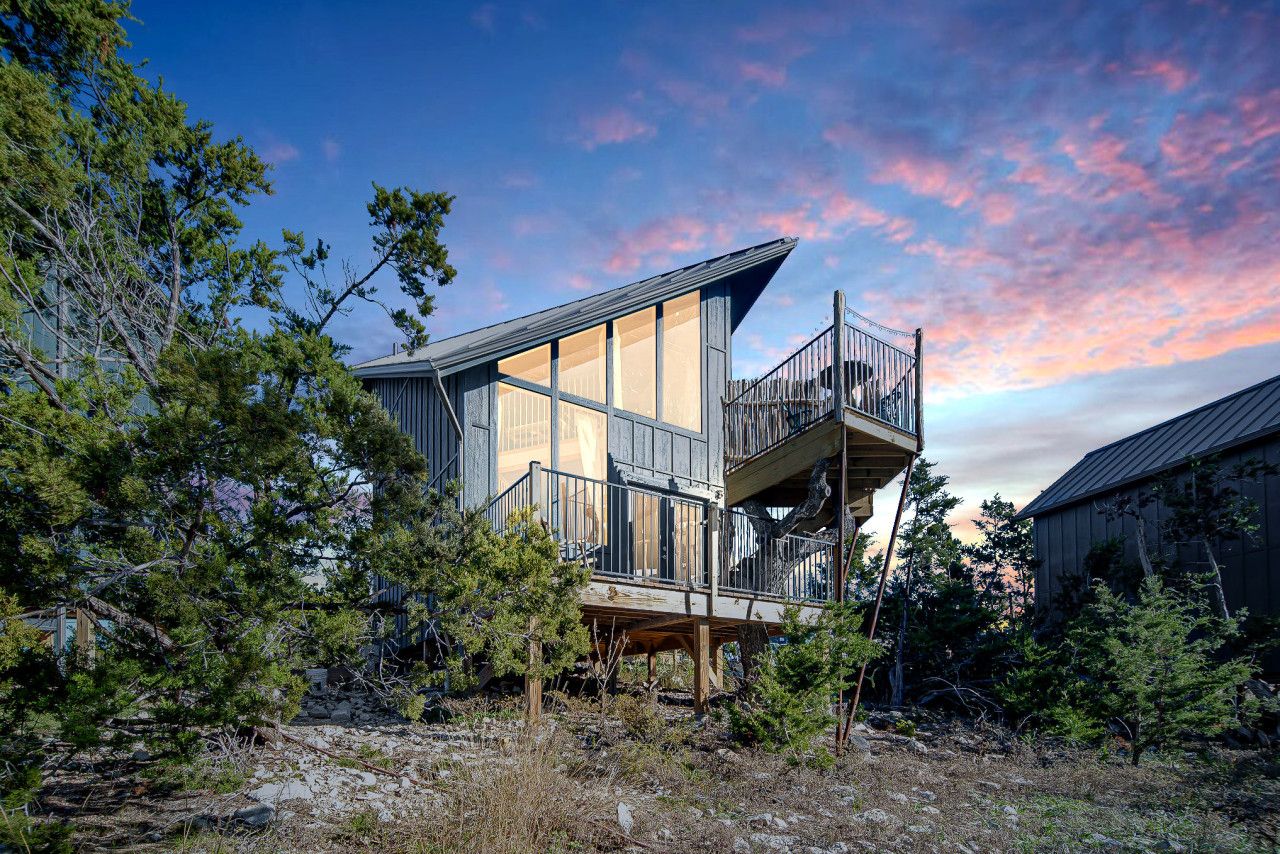 A modern two-story house with a balcony, surrounded by trees and a clear sky.