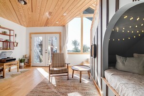 Un intérieur moderne avec un plafond en bois, un coin salon confortable avec un fauteuil et une petite table, un lit donnant sur un paysage à travers une grande fenêtre, et une bibliothèque remplie de divers objets.