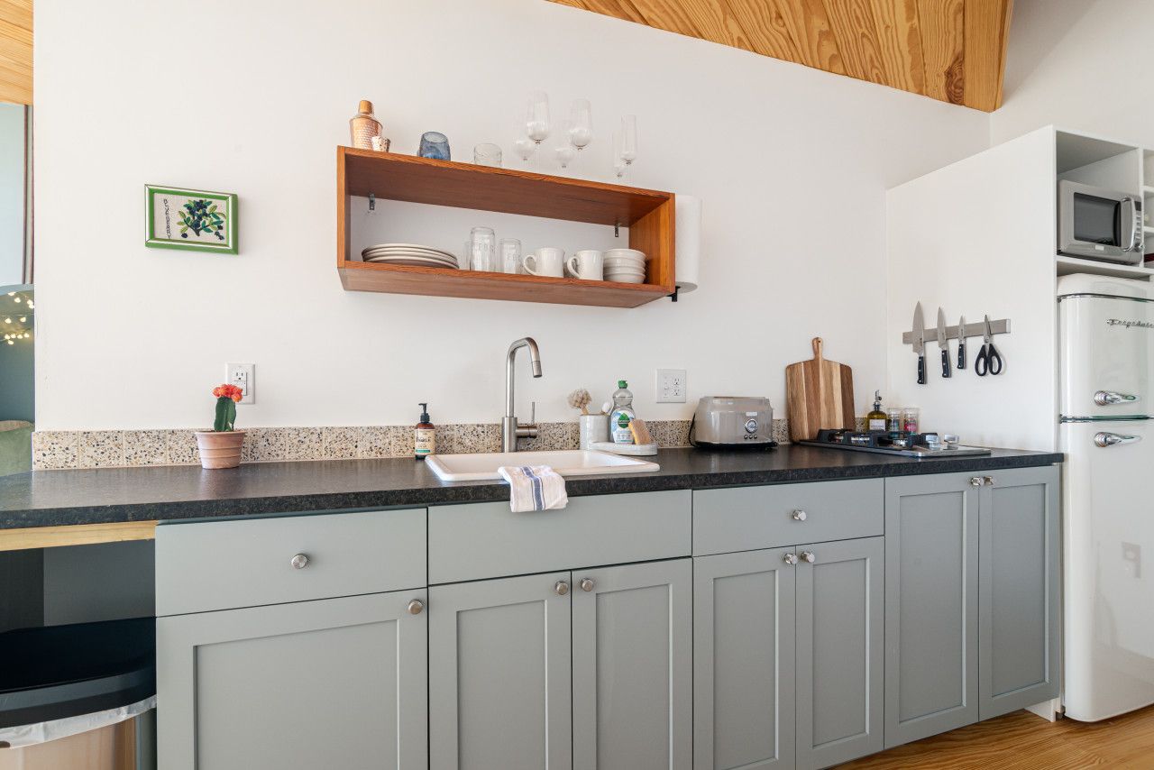 Une cuisine avec des meubles gris, un plan de travail noir, une étagère en bois au-dessus de l’évier et un réfrigérateur blanc.