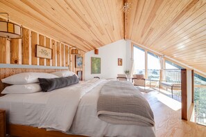 Une chambre avec un plafond en bois, un grand lit avec une literie blanche, une vue sur l’extérieur depuis un balcon et un tableau accroché au mur.
