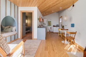 A cozy interior with a wooden ceiling, a seating area with a sofa and a chair, a kitchen with a dining table and chairs, and a hallway leading to another room.