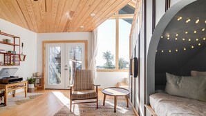 Un intérieur moderne avec un plafond en bois, un coin salon confortable avec un fauteuil et une petite table, un lit donnant sur un paysage à travers une grande fenêtre, et une bibliothèque remplie de divers objets.