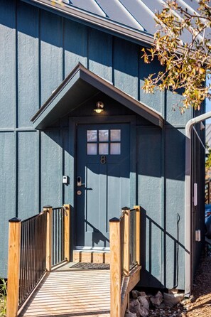 A building with a dark blue exterior, a wooden front porch with a black railing, and a door with a small window. - Blue Sage No. 8 - The Diagonal Treehome (Fredericksburg)