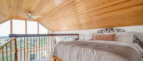 A bedroom with a wooden ceiling, a bed with white bedding, and a balcony with a railing.