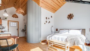 Une chambre avec un plafond en bois, un lit avec une literie blanche, un banc et un coin repas comprenant une table et des chaises.