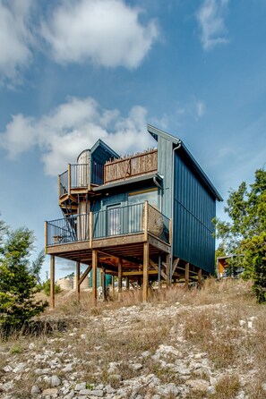 A two-story wooden structure with a balcony, situated on a rocky terrain with sparse vegetation.