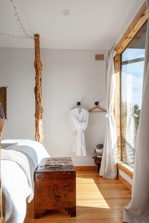 Un dormitorio con suelo de madera, una cama con ropa de cama blanca, un banco de madera, una bata colgada en un perchero de madera y una ventana con cortinas.