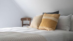 Una cama bien hecha con sábanas blancas y dos almohadones decorativos, una silla de madera y un techo inclinado.