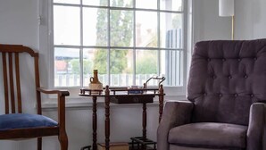 Una habitación con un ventanal grande, una silla de madera con cojín azul, un sillón púrpura y una pequeña mesita de madera con libros y un jarrón.