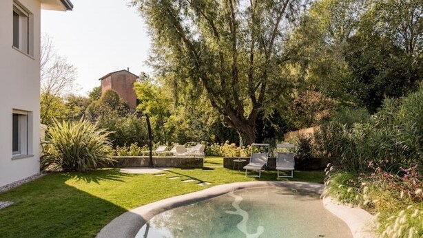 A swimming pool with a curved edge, surrounded by a well-maintained lawn and various plants. - CENTURY 1 APARTMENTS " SAN MARCO" (Peschiera del Garda)