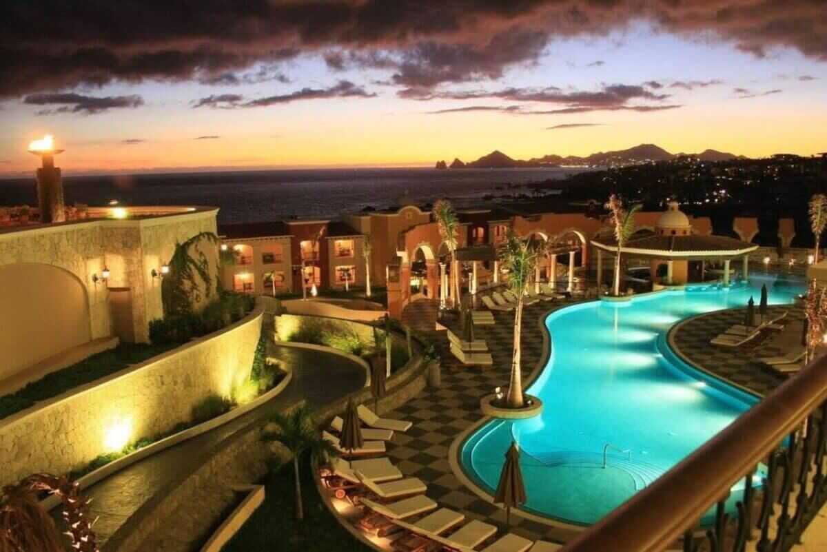A resort pool area with a view of the ocean at sunset, featuring a checkered pool deck, lounge chairs, and a small fountain.