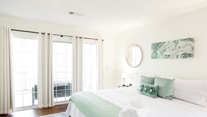 Een slaapkamer met een groot bed, een raam met gordijnen, een nachtkastje met een lamp en een spiegel aan de muur.