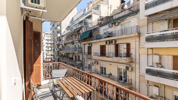 A balcony with a wooden table and chairs, overlooking a multi-story residential building with balconies and air conditioning units. - Vista: Spacious city centre home (Thessaloniki)