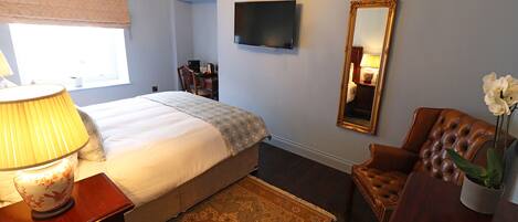 A bedroom with a bed, bedside table, lamp, TV, mirror, chair, and a potted plant.