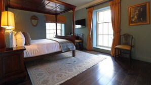 A traditional bedroom with a four-poster bed, a wooden nightstand, a desk, and a chair. There is a large window with curtains, a television on the wall, and a framed picture.