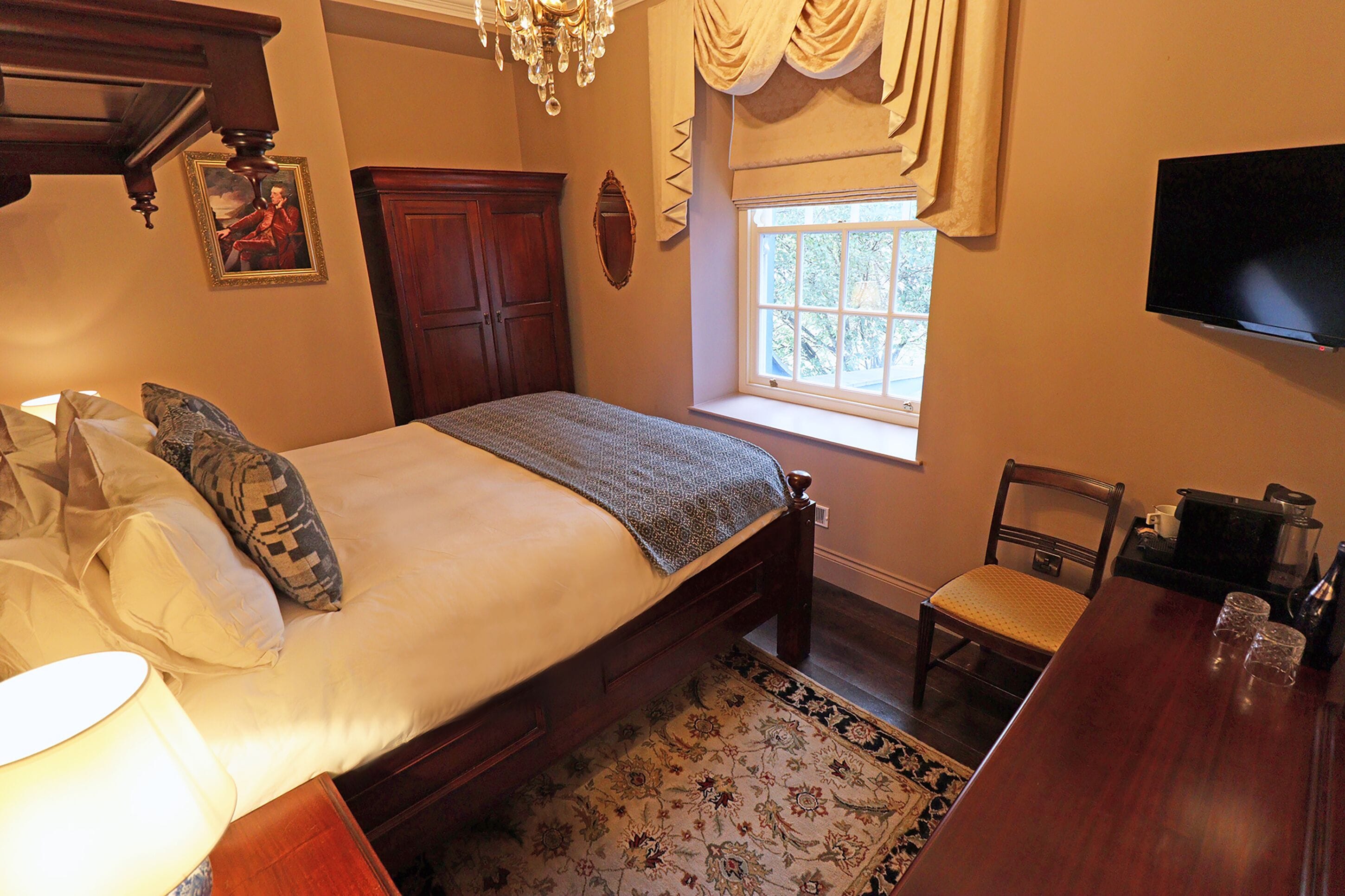 A bedroom with a bed, a wooden chair, a table with glasses, a TV, and a window with curtains.