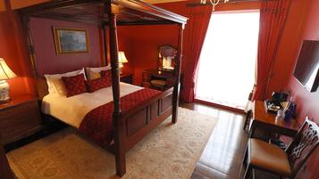 A four-poster bed with a red bedspread, a wooden nightstand with a lamp, a dresser, and a desk with a chair.
