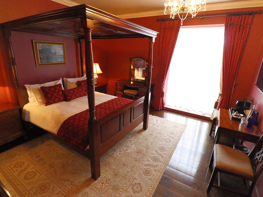 A four-poster bed with a red bedspread, a wooden nightstand with a lamp, a dresser, and a desk with a chair.