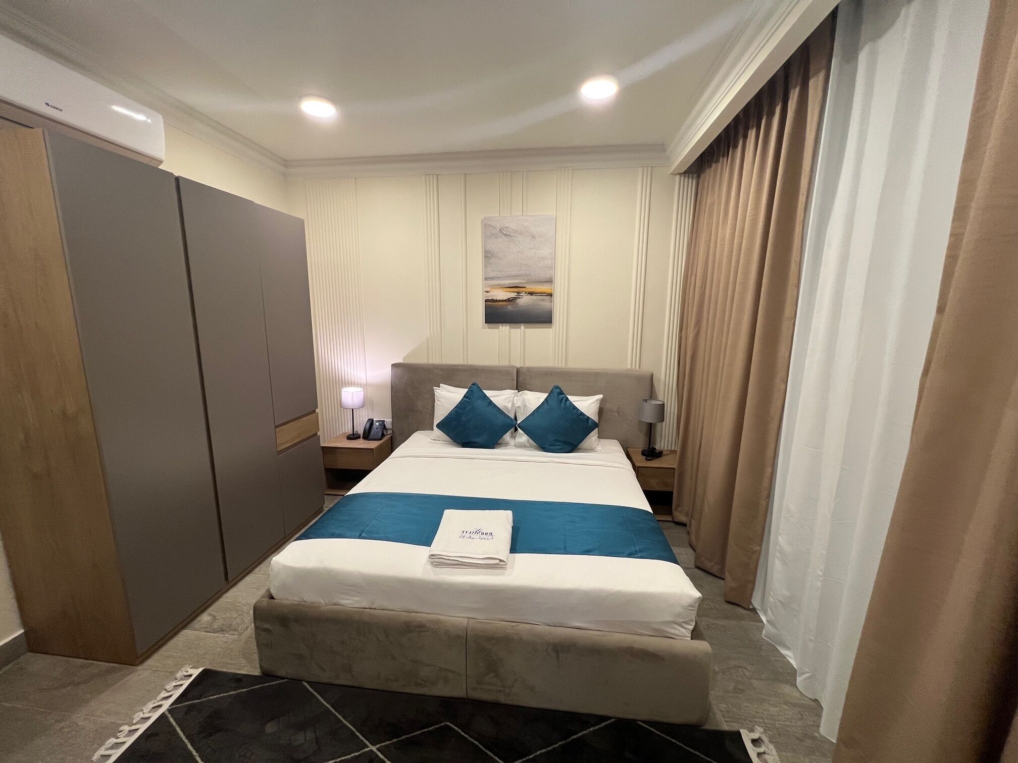 A hotel room with a bed, a nightstand, a lamp, a painting on the wall, and a wardrobe.