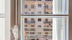 Vista a través de una ventana con barandilla de metal, mostrando un edificio de varios pisos con balcones y unidades de aire acondicionado.