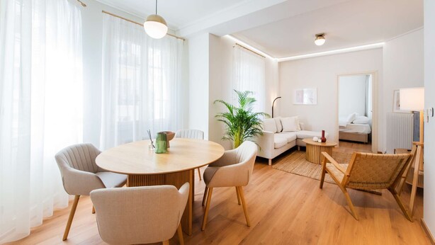 Una sala moderna con una mesa redonda de madera, sillas color beige, un sofá y una planta en maceta.
