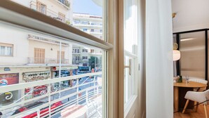Un balcon donnant sur une rue, un coin repas avec une table et des chaises, et une fenêtre offrant une vue sur des immeubles et des voitures garées.