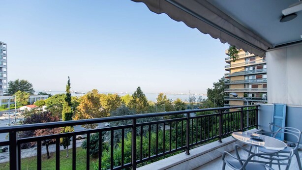 A balcony with a table and chairs, overlooking a cityscape with buildings and a body of water. - City Moon: Chic apt with park and sea view (Thessaloniki)