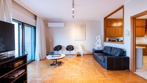 A modern living room with a wooden herringbone floor, a black leather sofa, a round coffee table, two black chairs, a TV on a stand, and a kitchen area visible through an open door. - City Moon: Chic apt with park and sea view (Thessaloniki)