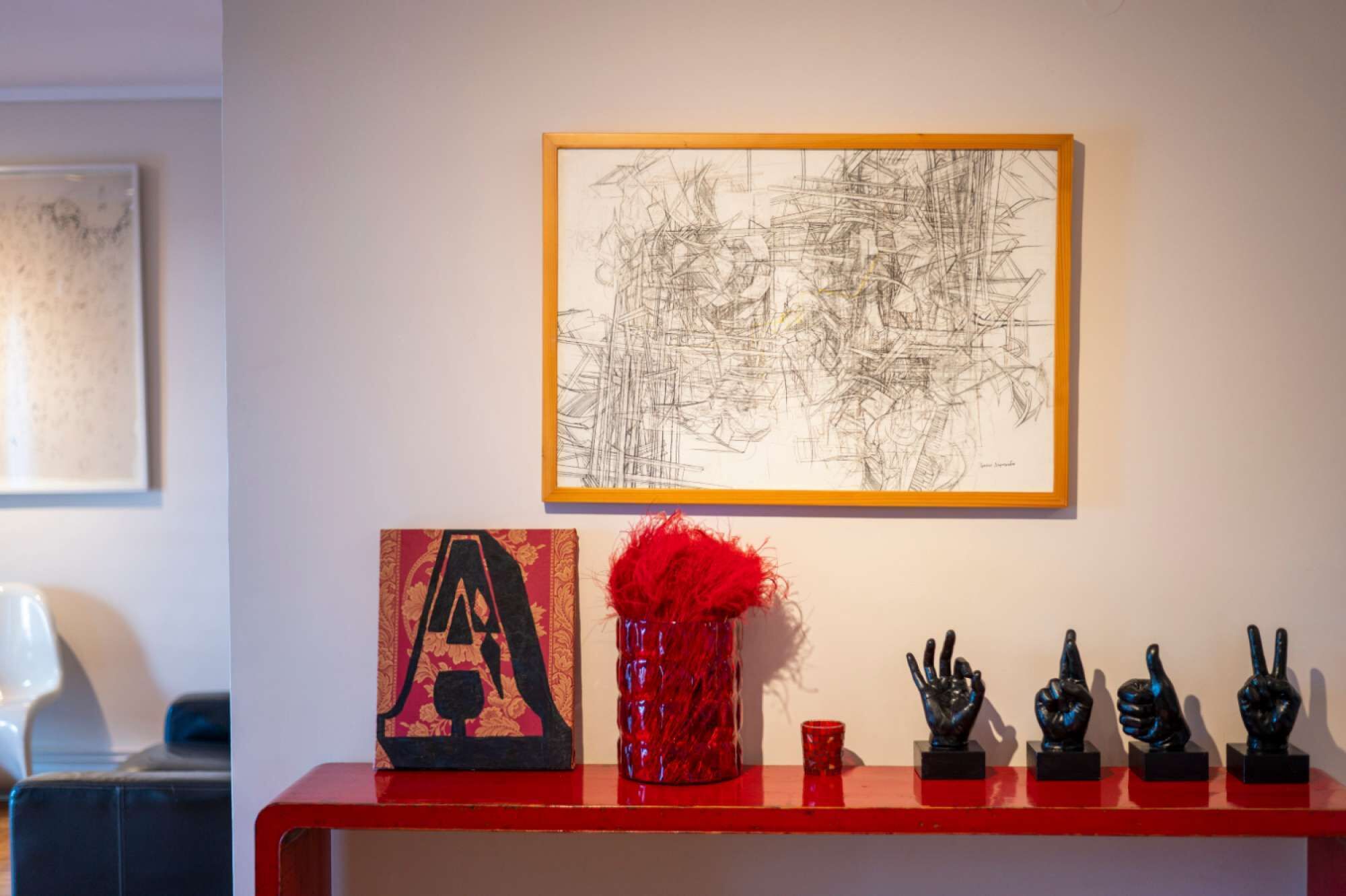 A red shelf with a framed abstract painting, a red vase with red feathers, and black sculptures of hands.