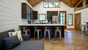 A modern kitchen with wooden ceiling beams, a central island with bar stools, and large windows. - Moonlight Ridge-Loft/Privacy/HotTub (Broken Bow)