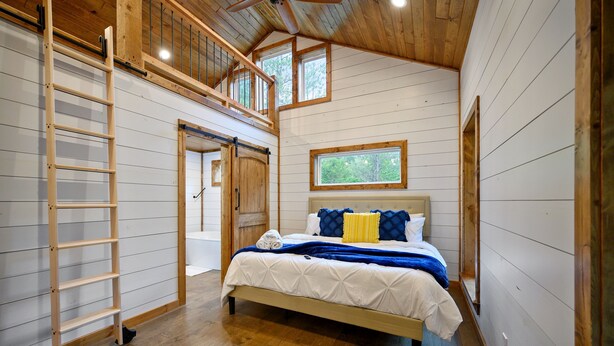 A bedroom with a loft area, a bed with blue and yellow bedding, a wooden ladder, and a window with a view of trees. - Moonlight Ridge-Loft/Privacy/HotTub (Broken Bow)