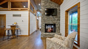 A cozy living room with a stone fireplace, wooden ceiling, and a large window. - Moonlight Ridge-Loft/Privacy/HotTub (Broken Bow)