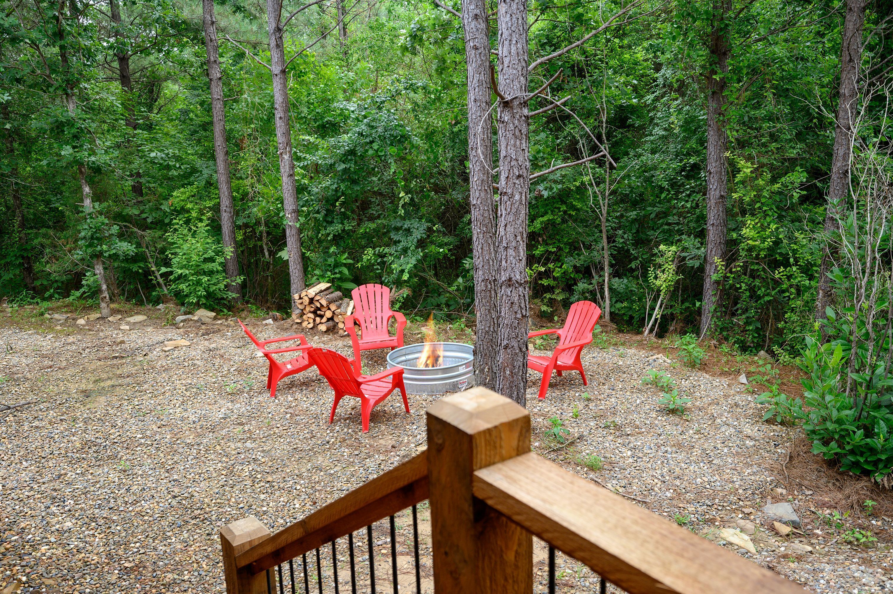 A forested area with a fire pit and red chairs.